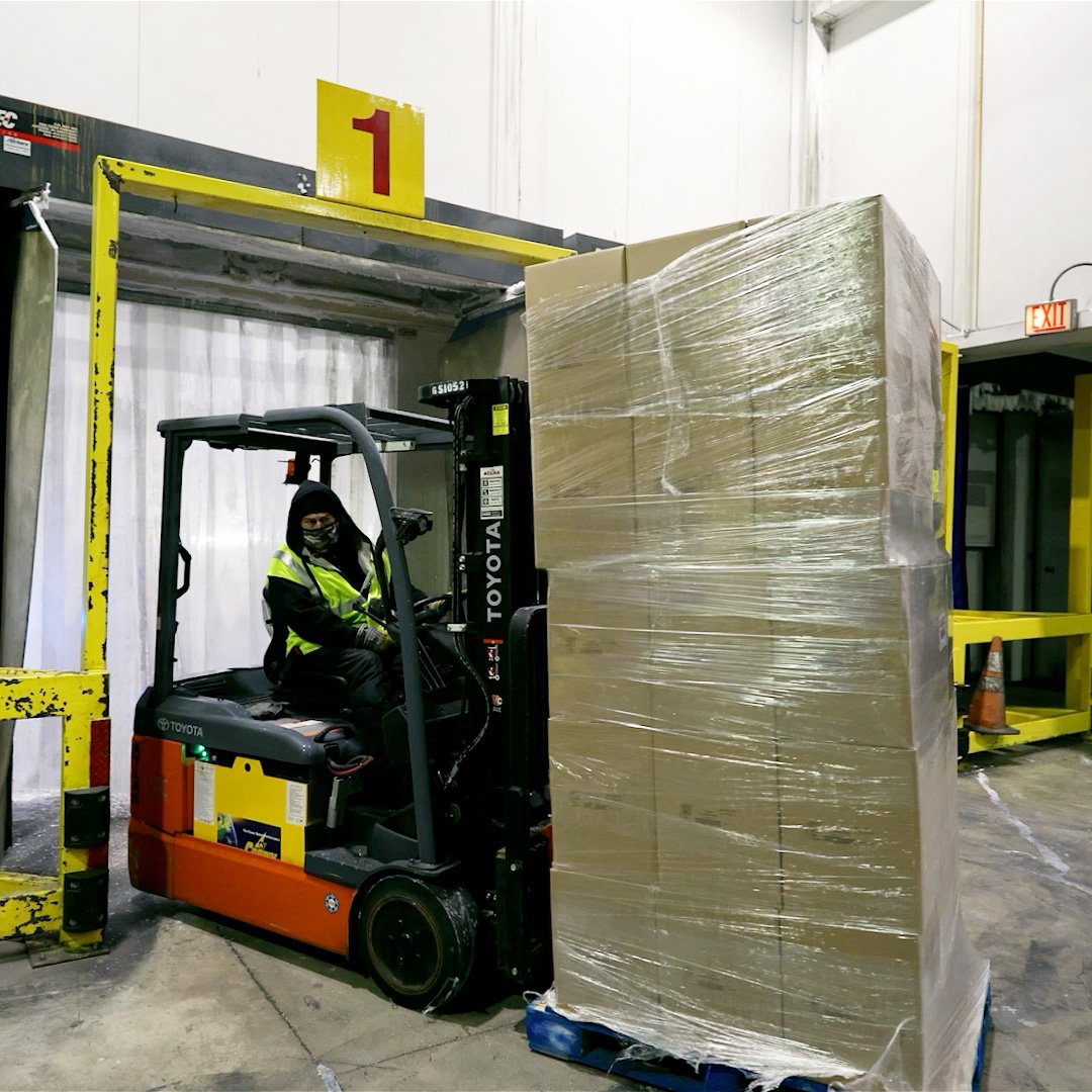 Forklift driver in protective gear moving a tall, wrapped pallet through a cold storage entry point.