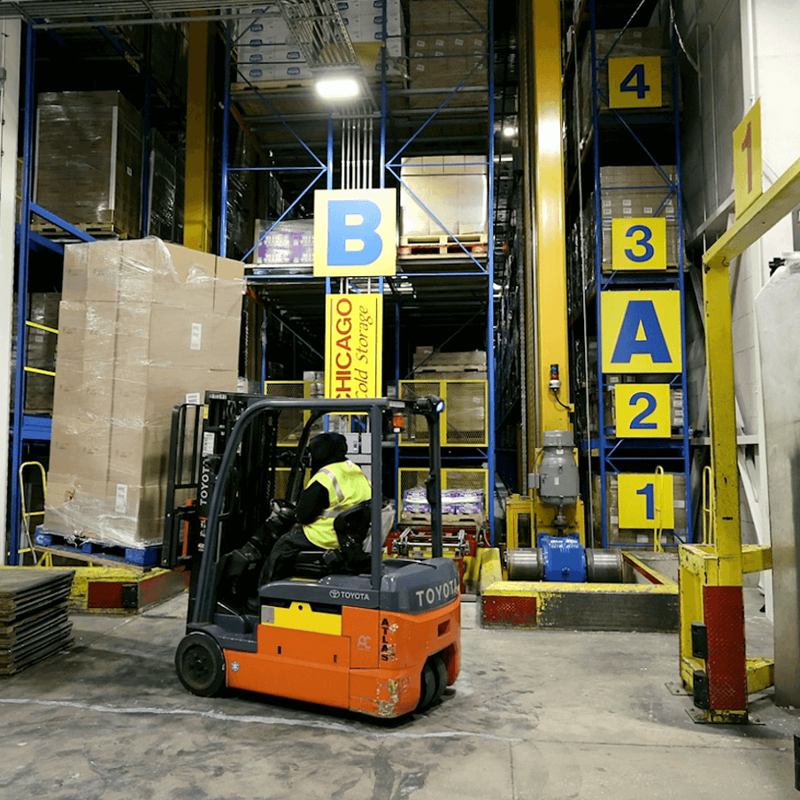 Forklift moving inventory inside a high-rack, labeled cold storage warehouse.