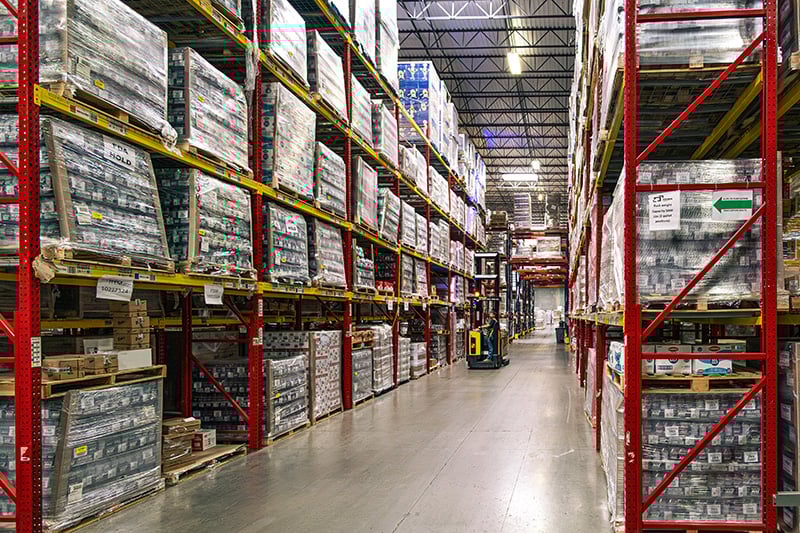 A warehouse worker operates a forklift in a wide aisle flanked by red and yellow high-rack storage systems.