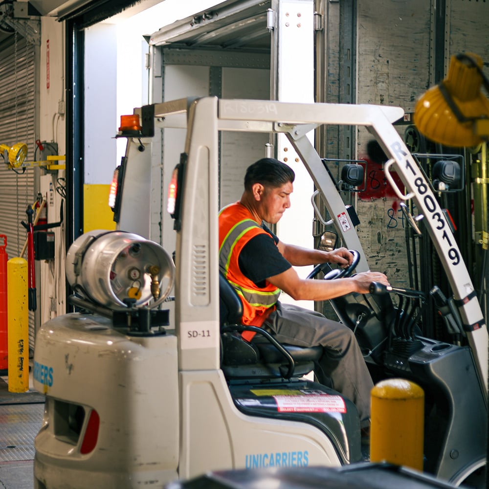 Forklift operator in a safety vest loading a semi-trailer at an indoor dock.