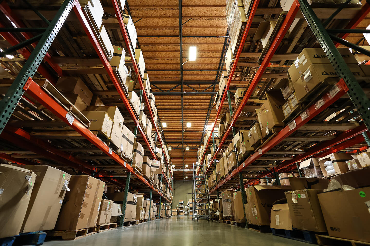 Low-angle view of a large warehouse aisle with tall storage racks and stacked cardboard boxes.