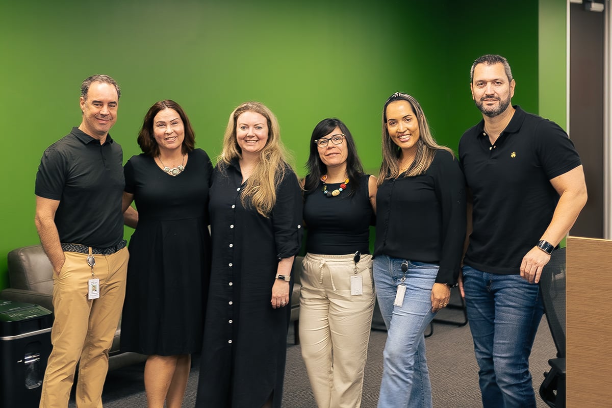 An group of six smiling professionals, the Source Logistics team, stands together in an office setting in front of a vibrant green wall.